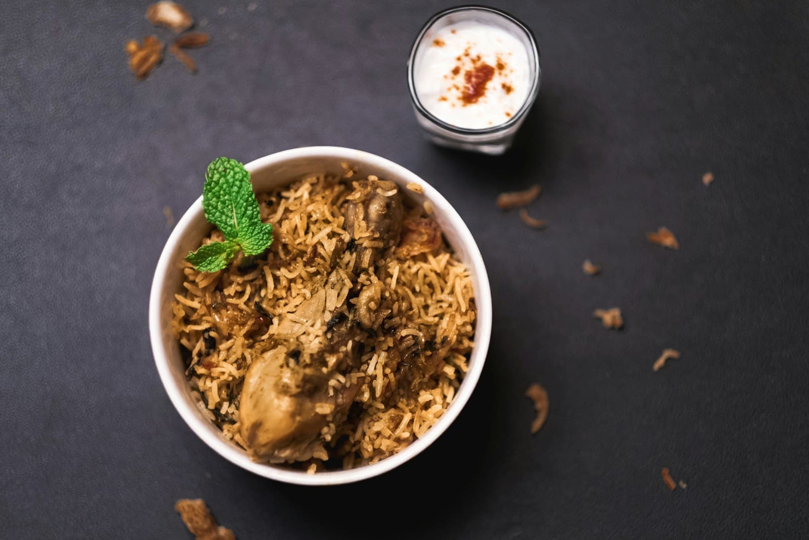 A bowl of mutton biriyani garnished with mint, with raita on the side